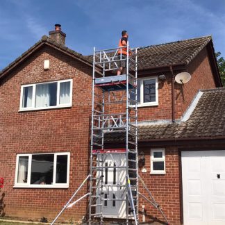 Worker stands on scaffolding, inspecting roof of a red-brick house with white-framed windows and garage door, under a clear blue sky.