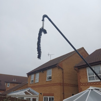 A long, curved chimney brush dangles from an extended pole against a cloudy sky, with two suburban brick houses featuring white conservatories in the background.