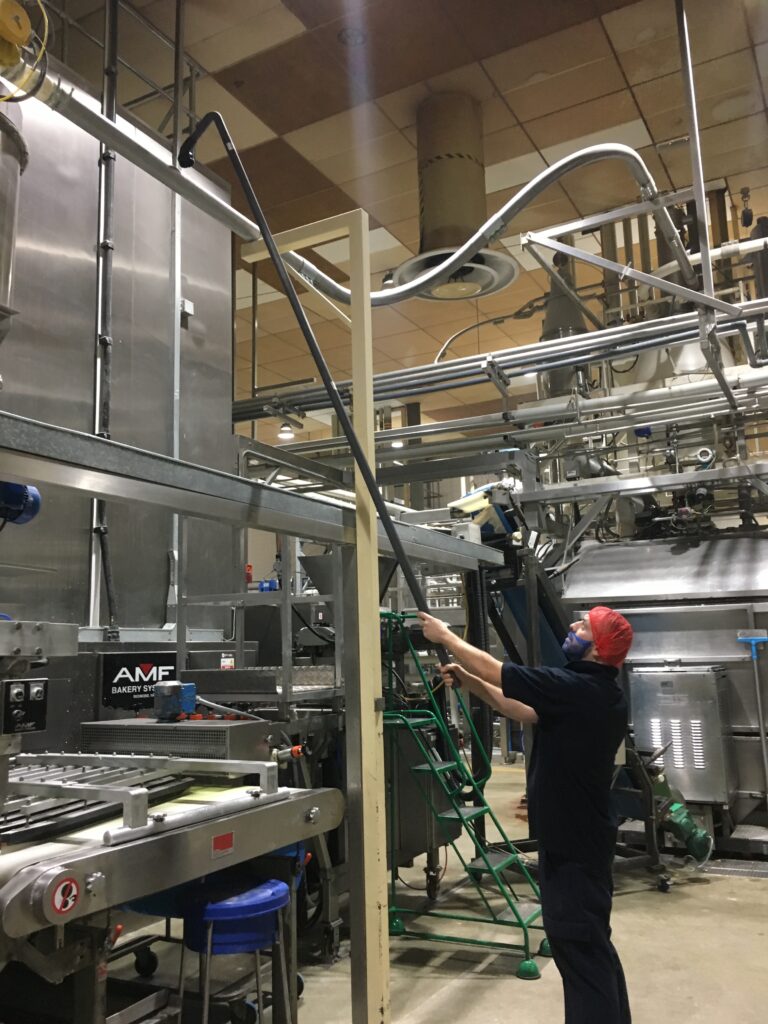 A worker wearing a red hairnet and mask uses a long pole to adjust equipment in an industrial bakery setting, surrounded by complex machinery labeled "AMF Bakery Systems."