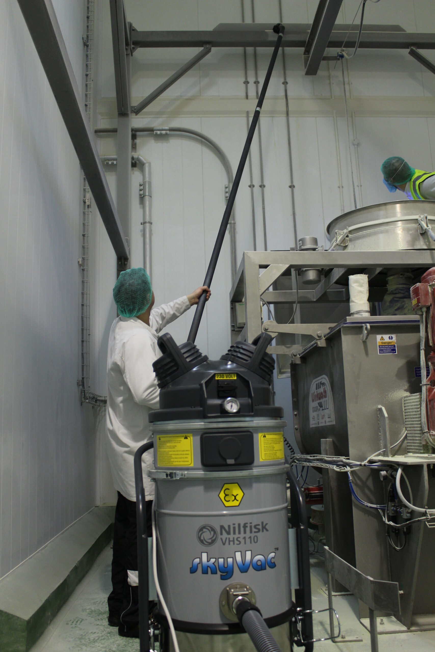 A person in protective gear uses a long pole attached to a Nilfisk VHS110 SkyVac machine for cleaning in an industrial setting with metal equipment and pipes.
