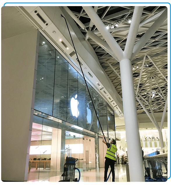 A person in a neon vest cleans high glass windows with a long pole inside a spacious modern shopping area, featuring an illuminated Apple logo.