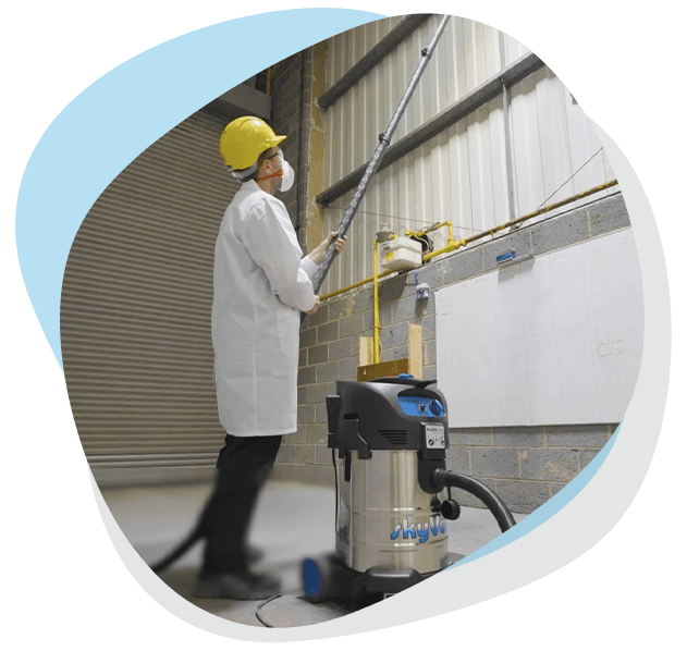 Worker wearing a yellow helmet and mask, using an industrial vacuum with a long nozzle to clean a high warehouse wall; "SkyVac" printed on the vacuum.